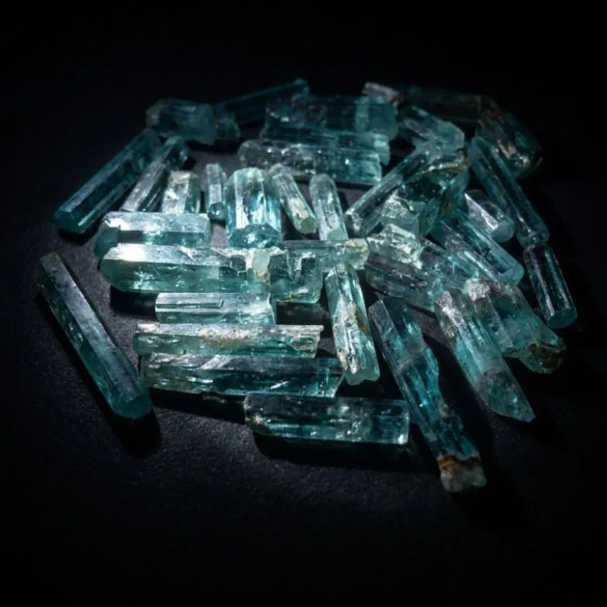 A parcel of rough tourmaline on a dark background, elongated blue-green prismatic crystals showing natural striations along their length